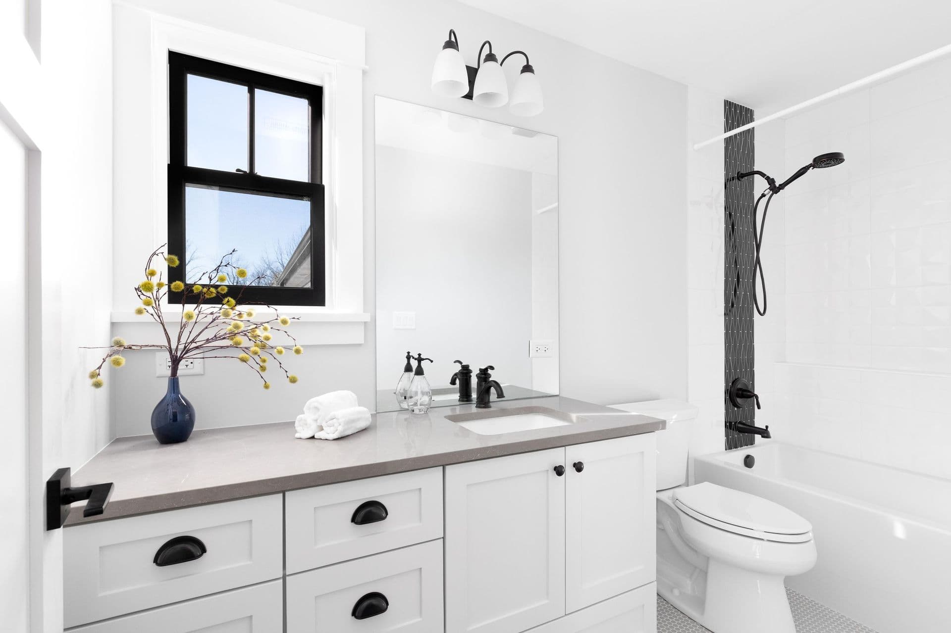 A white bathroom with white cabinets and gray countertop, black fixtures, and a shower with black tile accent.